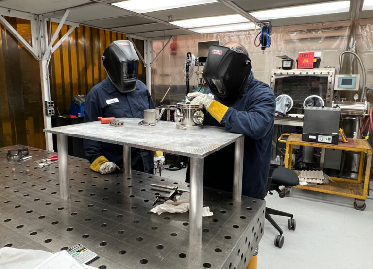 Two welders in protective gear work together on a fabricated chamber in a workshop