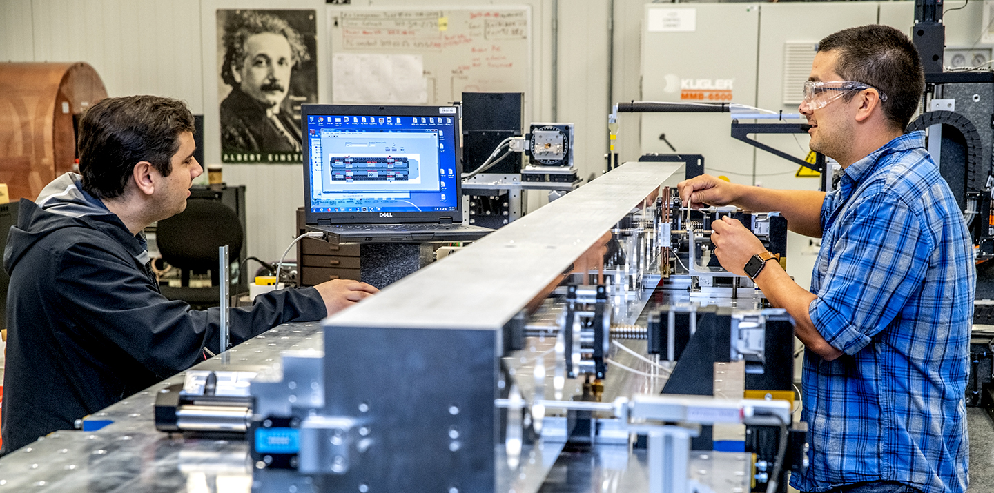 Two researchers working at a laboratory workbench with precision equipment, one gesturing while the other sits at a computer workstation.