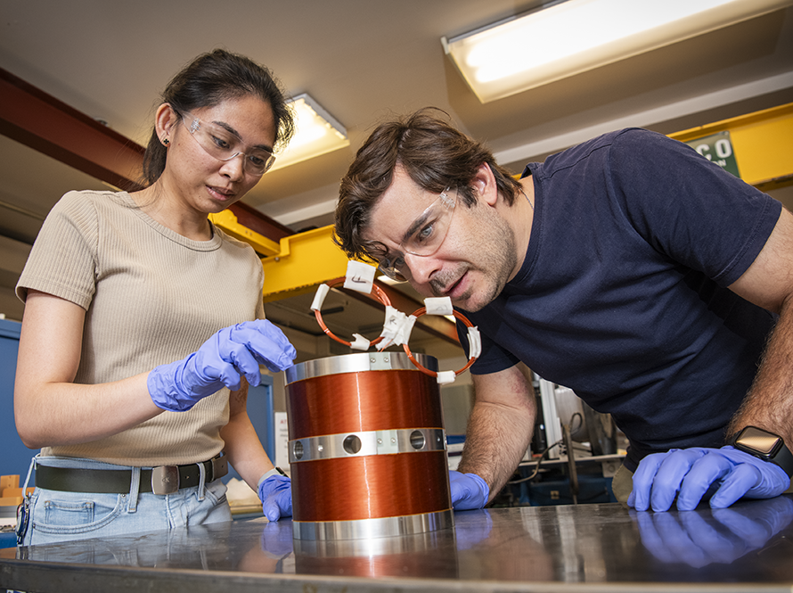 Two researchers wearing safety gloves examining a cylindrical metal component in a laboratory