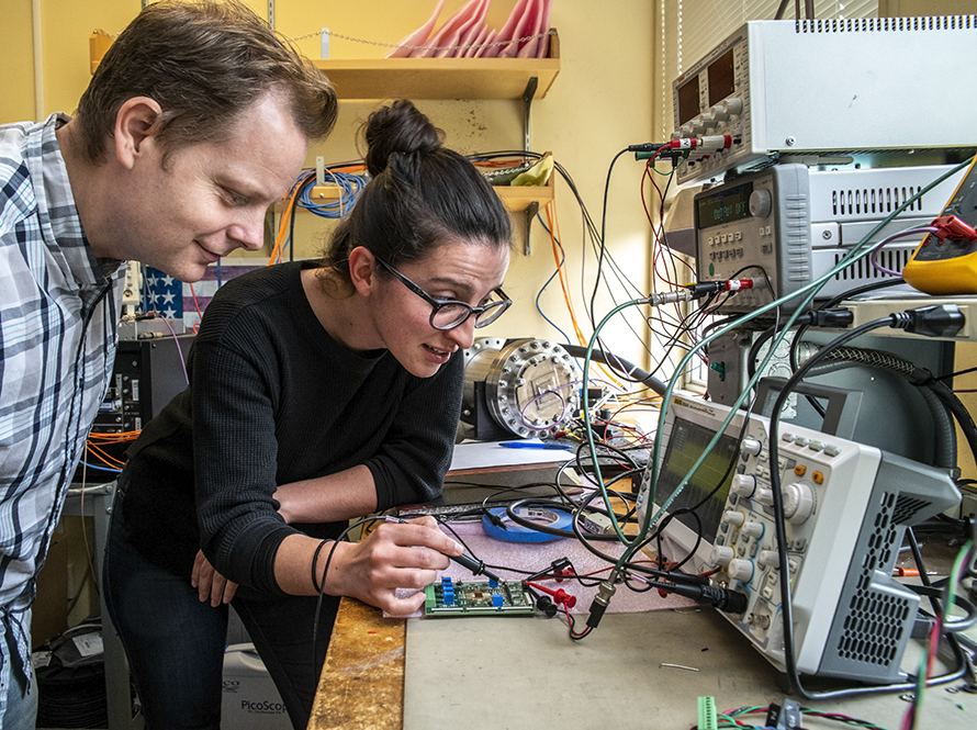 Two researchers working on an electronic circuit board at a lab bench, surrounded by testing equipment and connected wiring.