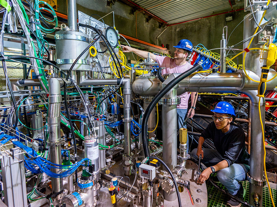 Two engineers in hard hats working on densely packed experimental equipment with pipes, gauges, and instrumentation.