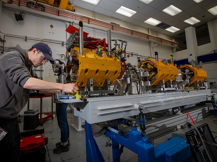 Engineer performs precision measurements on a series of yellow mechanical assemblies mounted on a long workbench.