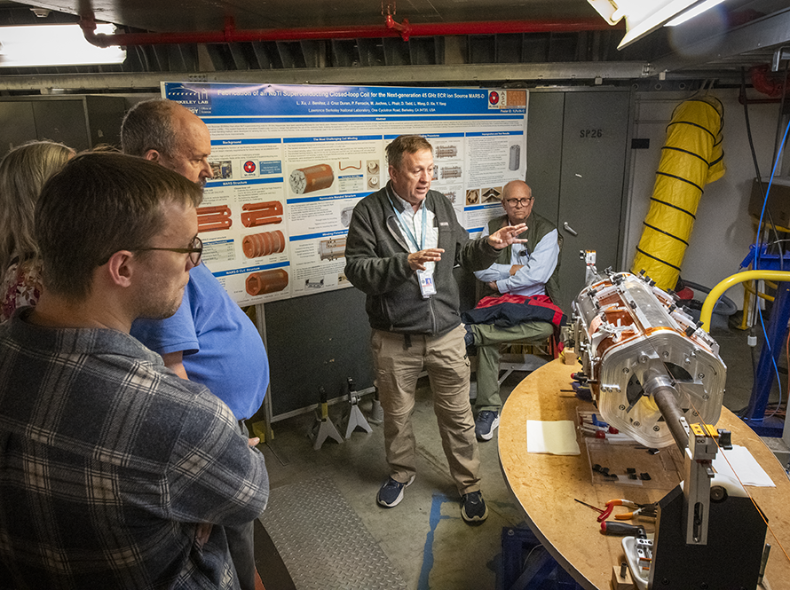 Presenter explaining a cylindrical magnet assembly to a group of colleagues in a laboratory, with a technical research poster behind them.