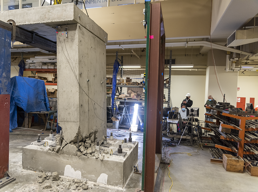 Close-up of fractured concrete support structure with technicians in the workspace behind.