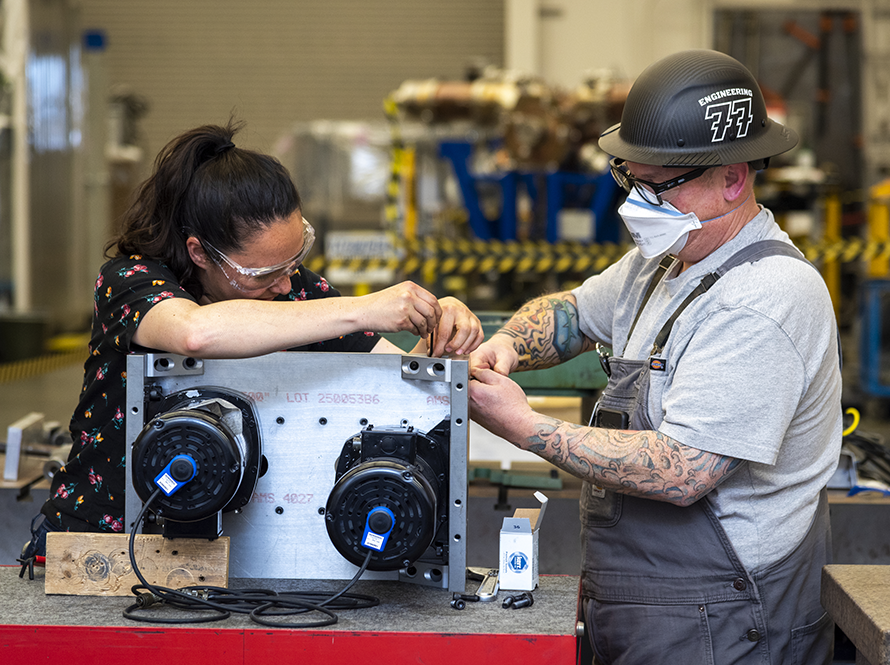 Two engineers wearing safety glasses and masks assemble components on a metal equipment housing.