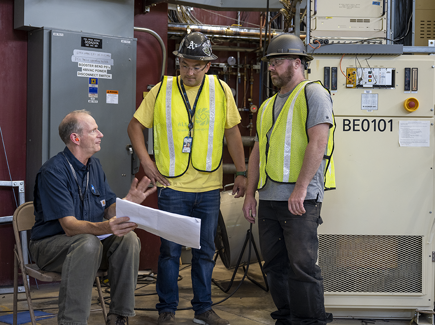 A lab staff member talks to two workers in safety vests and hard hats.