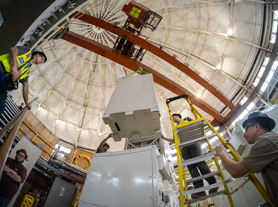 Wide-angle view of workers wearing hard hats working on equipment installation inside ALS with overhead crane.