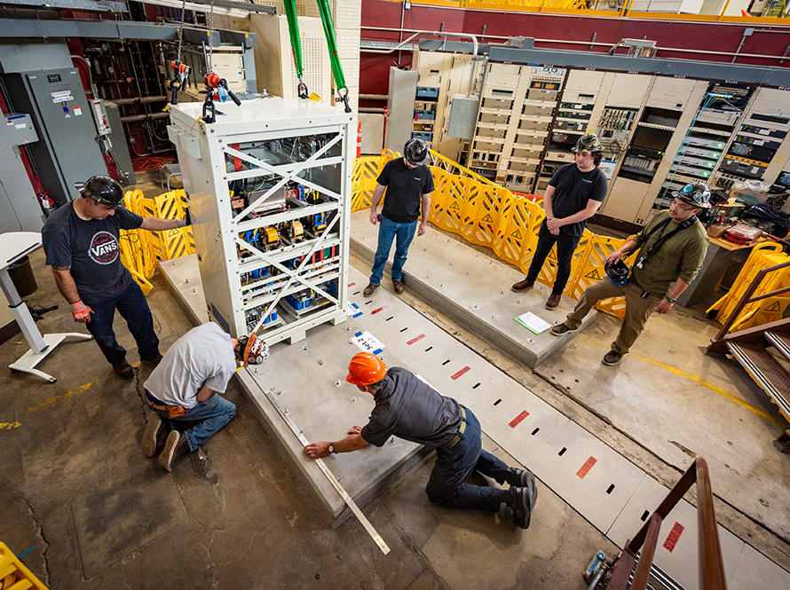 Group of technicians in hard hats positioning and securing a tall instrumented cabinet frame.