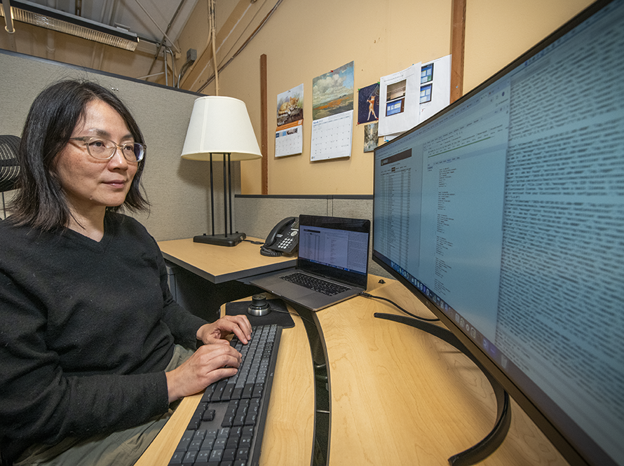 Researcher working at a desk with a laptop and monitor displaying information.