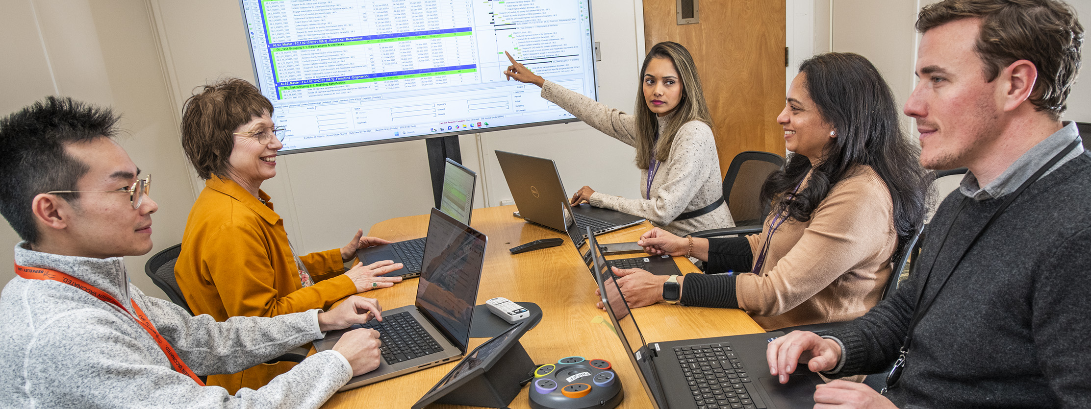 Team members in a meeting with laptops and one person pointing to the monitor.