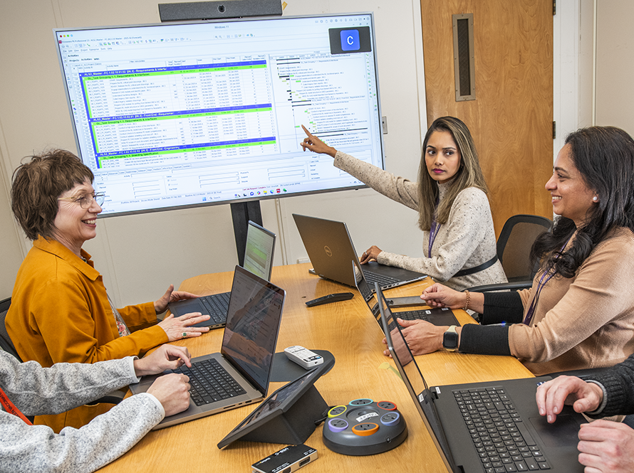 People with laptops in a conference meeting with one pointing to a monitor display.
