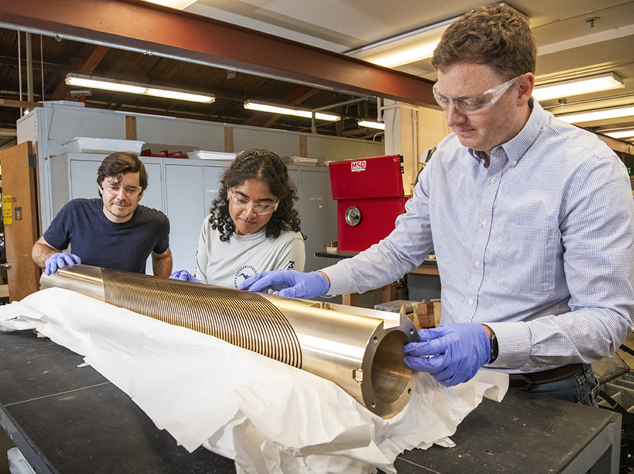 Three engineers in safety glasses inspect a cylindrical metal component.