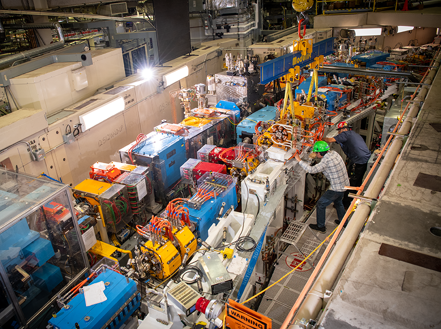 Overhead view of particle accelerator equipment under construction stretching through a laboratory space, with two workers in hard hats.