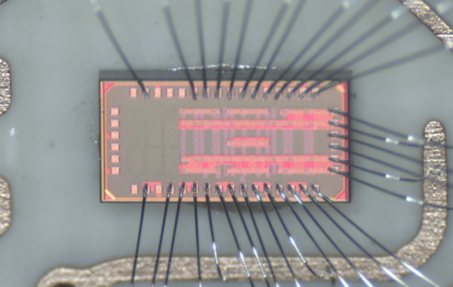 Magnified view of a rectangular microchip with fine wires bonded around its edges for electrical connections.